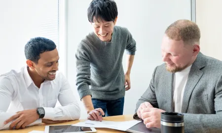 Three smiling male students stand around a table with documents in a classroom