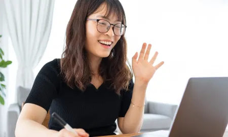 A young woman wearing glasses smiles and waves into a laptop camera while taking notes during a virtual class