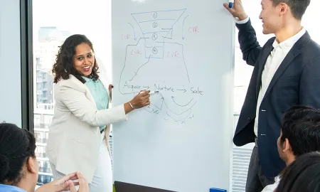 Two MBA students present a sales funnel diagram on a whiteboard to their peers in a bright classroom.