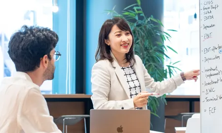 A smiling student in a white blazer points to a whiteboard listing financial terms like EBITDA and FCF while a student at the same table listens.