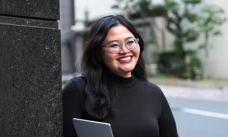 A smiling woman with glasses and long dark hair, wearing a black turtleneck and holding a tablet outdoors.
