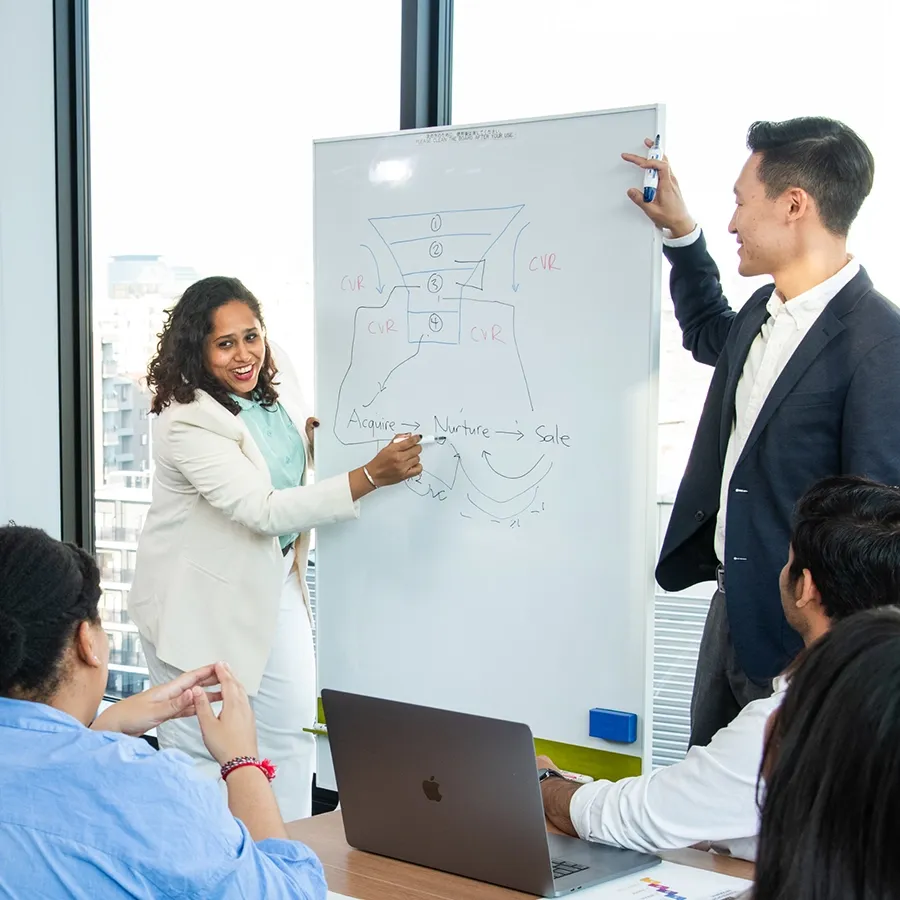 Two MBA students present a sales funnel diagram on a whiteboard to their peers in a bright classroom.