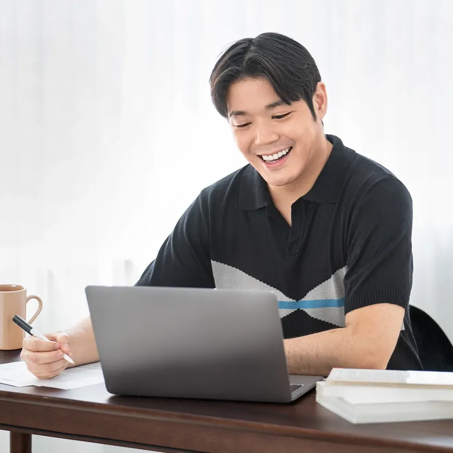 A student smiling at a laptop while taking notes at a desk with books and a coffee mug nearby.