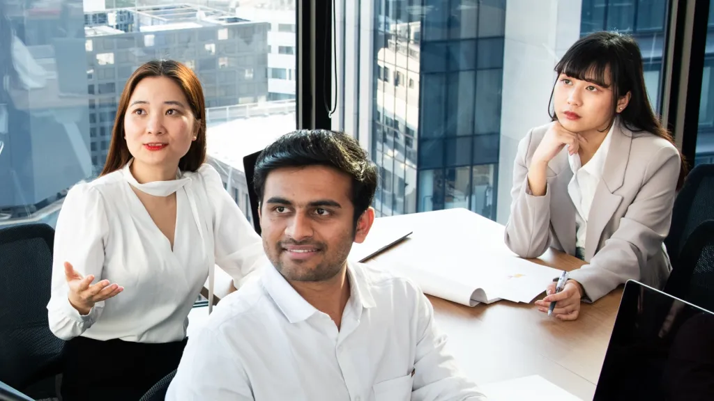 Three young professionals sitting at a conference table in a high-rise classroom, engaged in a collaborative group discussion.