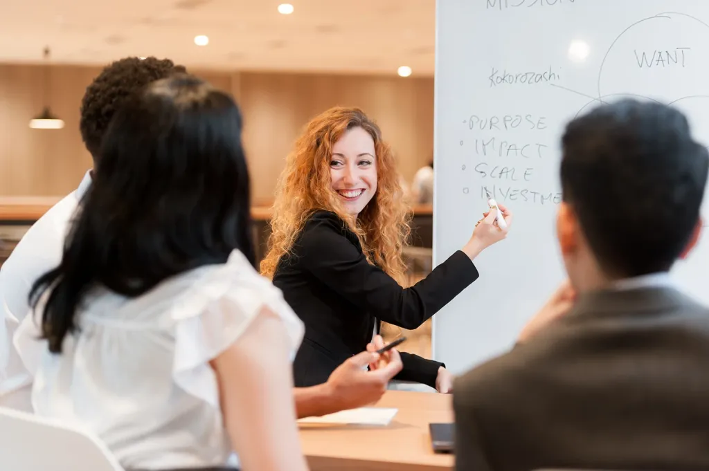 A smiling female MBA student leads a collaborative group discussion with classmates, writing on a whiteboard.